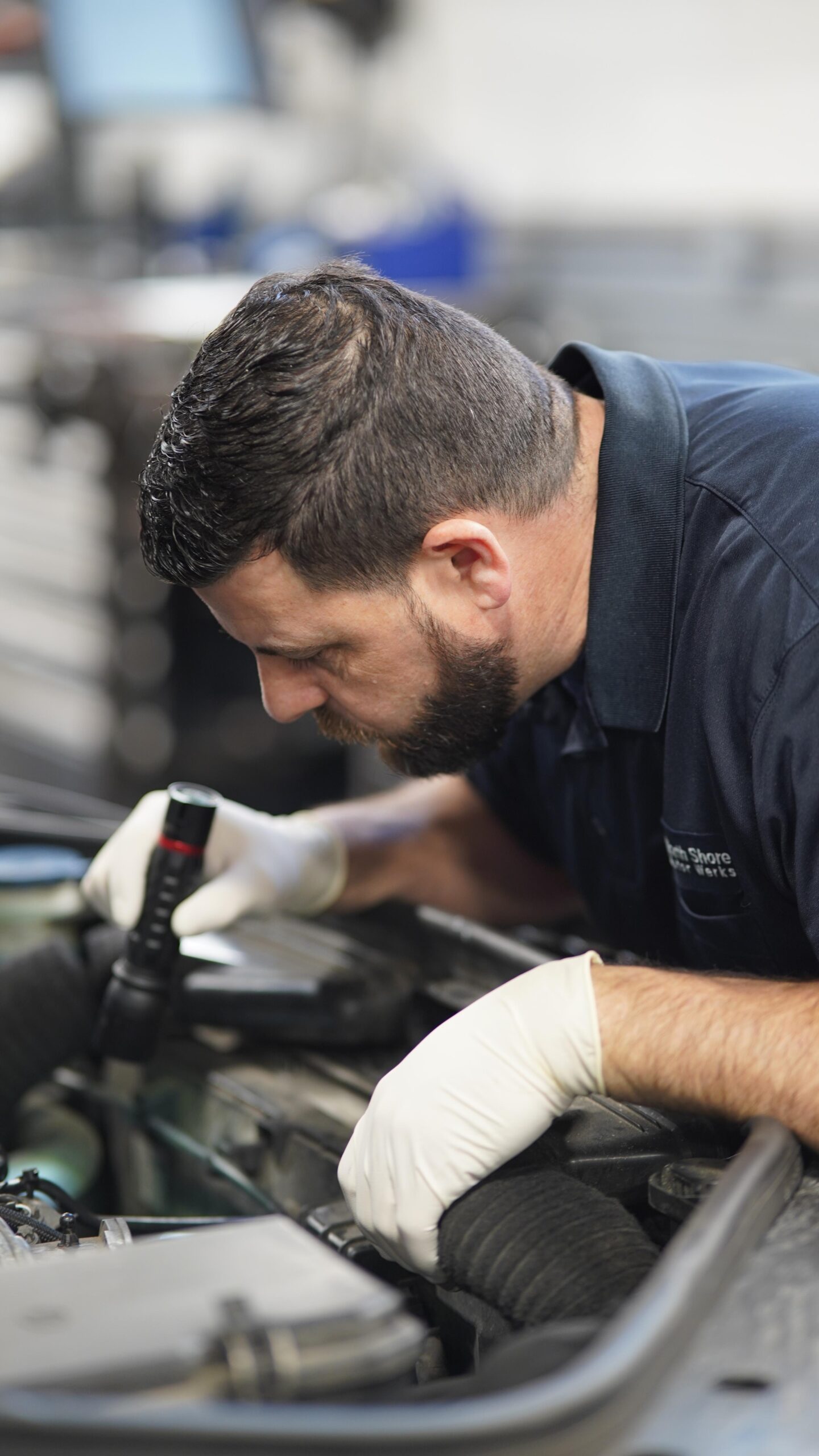 A mechanic with gloves conducts MINI maintenance by inspecting the car engine with a flashlight in the garage specializing in European auto Car Service.