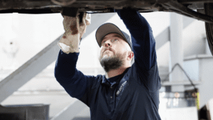 A mechanic in a navy shirt and cap uses a rag while inspecting or repairing the underside of a vehicle in a garage. He is performing a BMW oil change at North Shore Motor Werks.