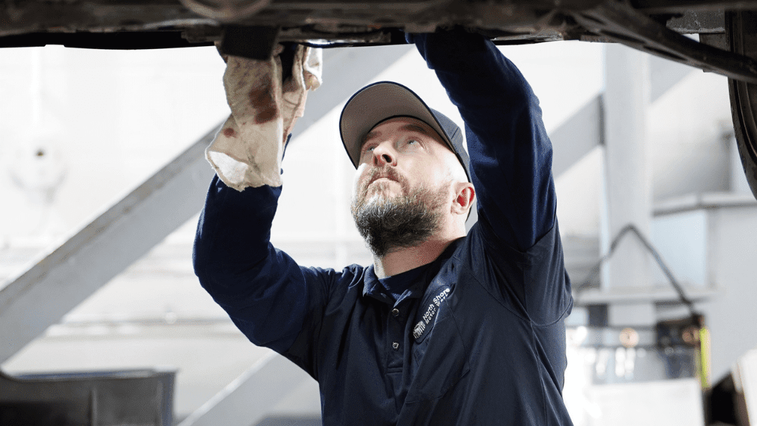 A mechanic in a navy shirt and cap uses a rag while inspecting or repairing the underside of a vehicle in a garage.