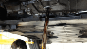 Oil is draining from the underside of a vehicle during an oil change, with the car lifted and a person partially visible below.