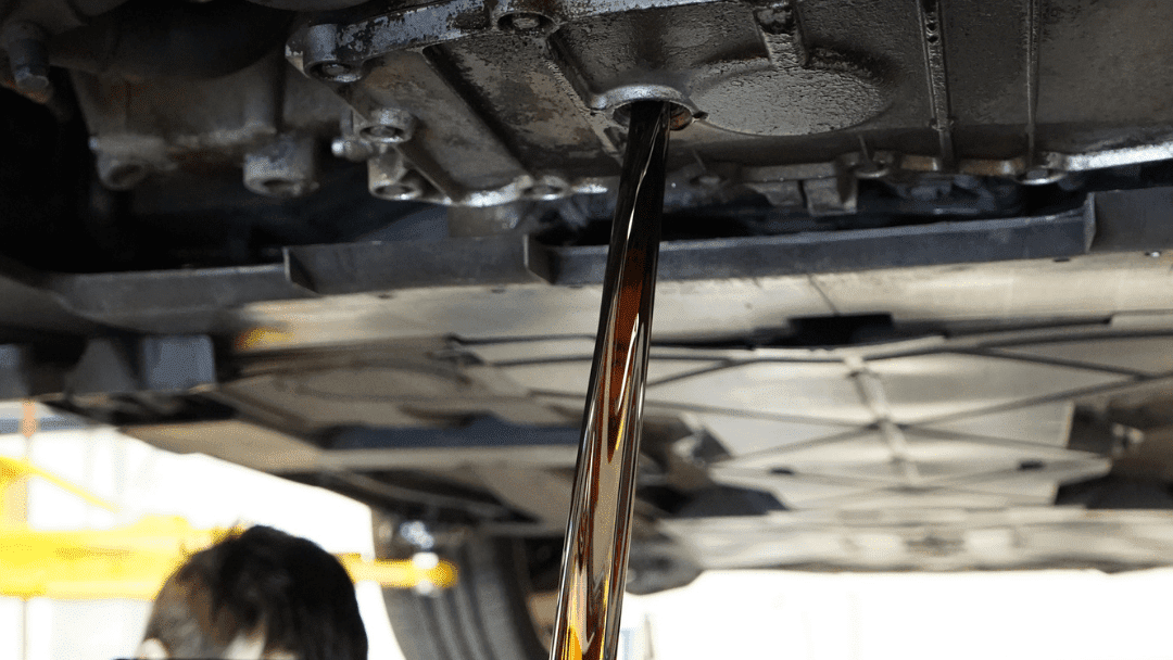 Oil is draining from the underside of a vehicle during an oil change, with the car lifted and a person partially visible below.