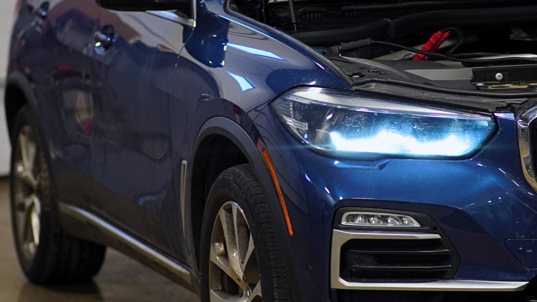 Close-up of the front left side of a blue SUV with headlights on and the hood partially open, parked indoors.