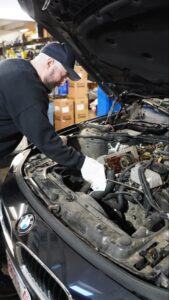 A mechanic wearing a cap and gloves works on the engine of a black BMW car with the hood open inside an auto repair shop.