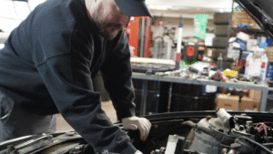 A mechanic wearing a black cap and sweatshirt inspects an open car engine in a workshop with various tools and tires in the background.