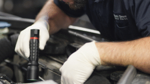 A mechanic wearing white gloves examines a car engine with a black flashlight, leaning over the engine bay.