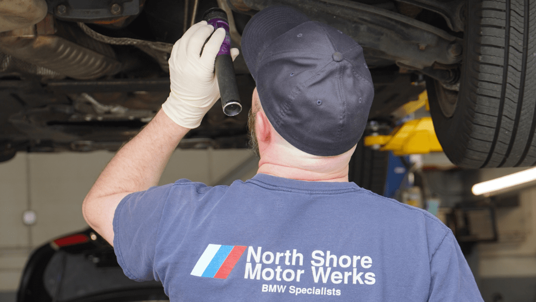 A mechanic wearing gloves and a "North Shore Motor Werks" shirt inspects the underside of a vehicle using a flashlight in an auto repair shop.