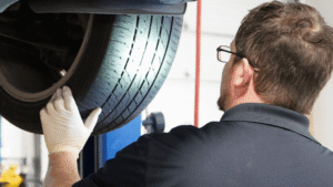 A mechanic wearing gloves inspects the tread on a car tire, which is raised on a lift in a garage.