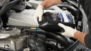 A person wearing gloves pours blue antifreeze coolant from a black container into a car engine’s coolant reservoir.