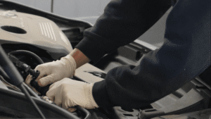 A person wearing white gloves works on a car engine, using both hands to handle components under the hood.