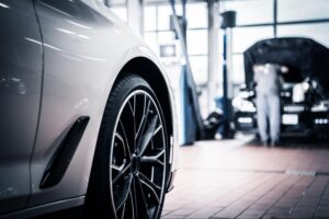 Close-up of a car wheel in a garage, with another car in the background being serviced by a mechanic.