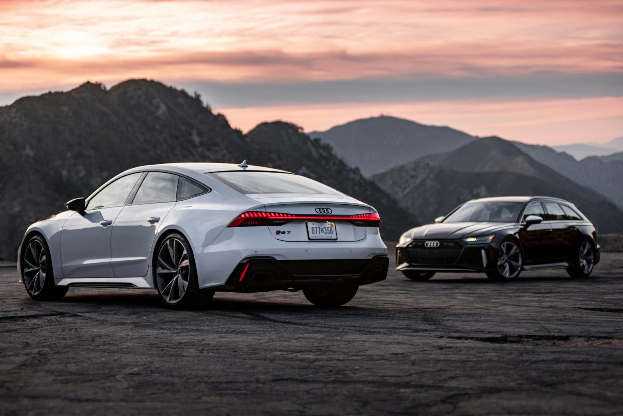 Two Audi cars are parked on an open road with mountains in the background at sunset, one a white sedan and the other a dark station wagon.