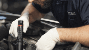 A mechanic wearing gloves inspects a car engine with a flashlight.
