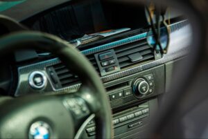 Close-up of a BMW car's dashboard, showing the steering wheel, air vents, climate control buttons, and a hanging accessory.