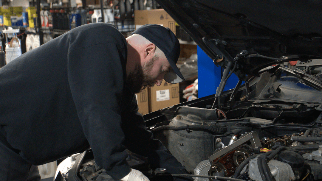 A mechanic in a black cap and sweatshirt works on the engine of a car in a garage with shelves and boxes in the background.